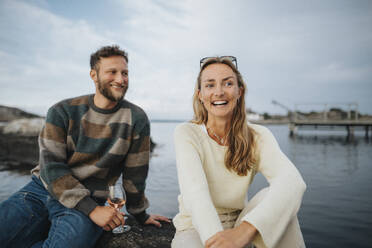 Glückliche Frau sitzt mit männlichem Freund und trinkt Wein, während sie auf einem Felsen in der Nähe eines Sees sitzt - MASF39234
