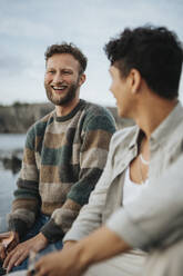 Glücklicher junger Mann genießt mit männlichen Freund in der Nähe von See sitzen - MASF39238