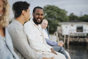 Smiling young man talking with male friend while sitting on pier near lake - MASF39240