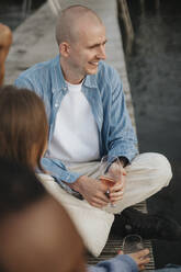 Lächelnder Mann hält ein Weinglas und sitzt im Schneidersitz auf einem Pier - MASF39241