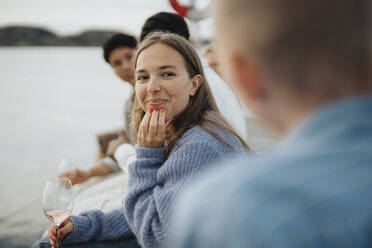 Lächelnde junge Frau mit Hand am Kinn, die mit Freunden auf einem Steg am See sitzt - MASF39247