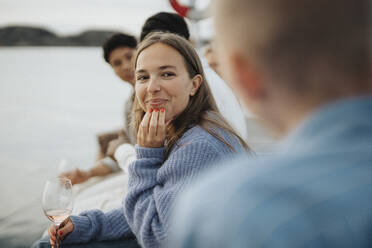 Smiling young woman with hand on chin sitting with friends on pier near lake - MASF39247