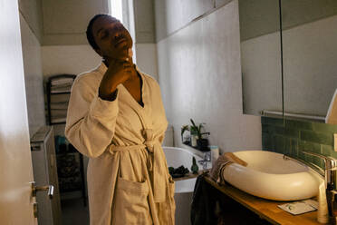 Young woman wearing bathrobe using jade roller on face in bathroom - MASF39273