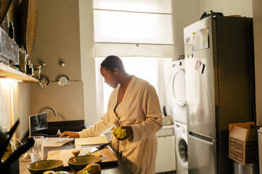 Young woman holding grapes using laptop in kitchen at home - MASF39279