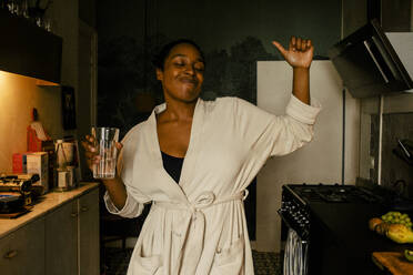 Carefree young woman holding drinking glass while dancing in kitchen at home - MASF39283