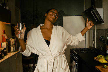 Carefree young woman holding drinking glass while dancing in kitchen at home - MASF39284