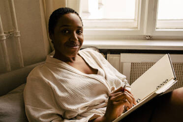 Portrait of smiling young woman with book in bedroom at home - MASF39291