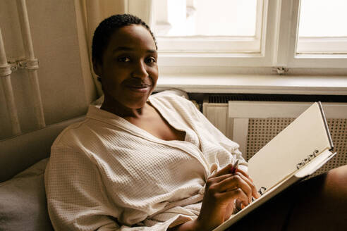 Porträt einer lächelnden jungen Frau mit Buch im Schlafzimmer zu Hause - MASF39291