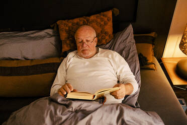 Senior man reading book while lying on bed at home - MASF39294