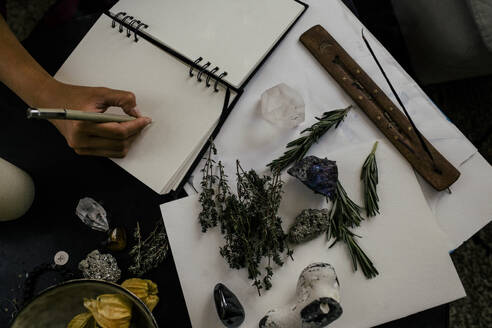 Ausgeschnittenes Bild einer Frau, die neben Kräutern und Kristallen auf einem Tisch in ein Buch schreibt - MASF39330