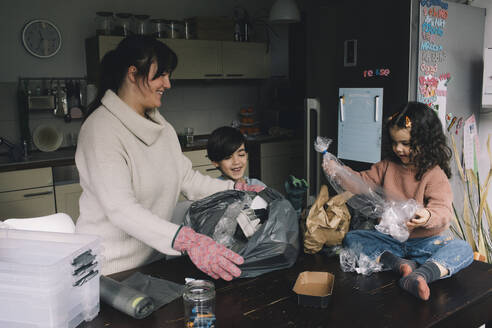 Smiling mother with son and daughter collecting plastic bags at home - MASF39353