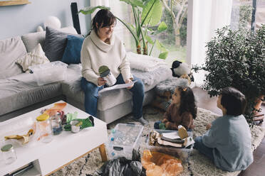 Smiling woman with son and daughter separating waste while sitting in living room - MASF39359