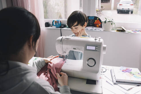 Son looking at mother sewing clothes at home - MASF39371