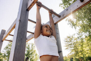 Low angle view of smiling girl hanging while doing monkey bars at summer camp - MASF39392