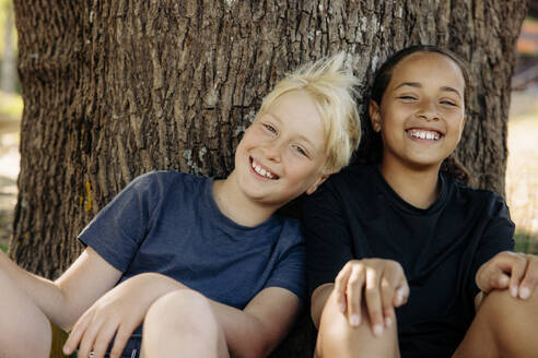 Portrait of happy boy sitting with female friend by tree trunk at summer camp - MASF39413