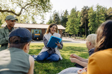 Happy female counselor doing activities with kids while sitting on grass at summer camp - MASF39422