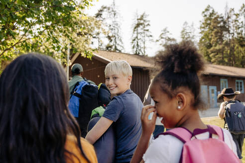 Portrait of smiling boy looking over shoulder while walking with friends at summer camp - MASF39427