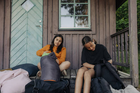 Mädchen packen Rucksäcke aus, während sie auf der Veranda vor der Hütte sitzen - MASF39436