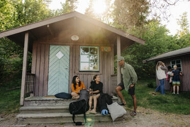 Männlicher Betreuer im Gespräch mit Mädchen, die auf der Veranda vor der Hütte im Sommercamp sitzen - MASF39438