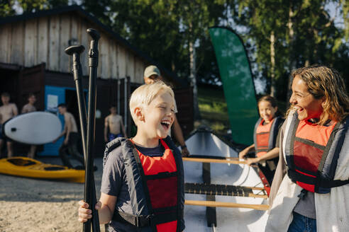 Glücklicher Junge mit Betreuer, der Schwimmwesten trägt, während er im Sommerlager Kajak fährt - MASF39448