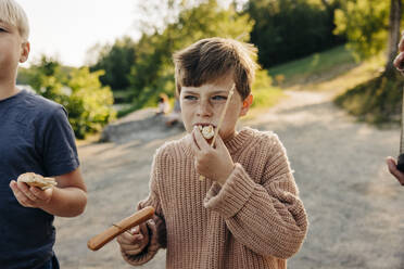 Boy wearing sweater and eating sausage with friend at summer camp - MASF39480