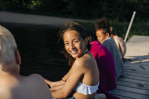 Seitenansicht Porträt von lächelnden Mädchen sitzen mit Freunden auf Steg in der Nähe von See im Sommerlager - MASF39491