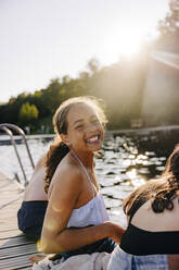 Side view portrait of happy girl laughing while sitting with friends on jetty near lake - MASF39495