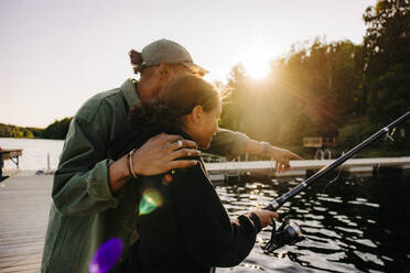 Male counselor gesturing by girl fishing on jetty near lake at summer camp - MASF39502