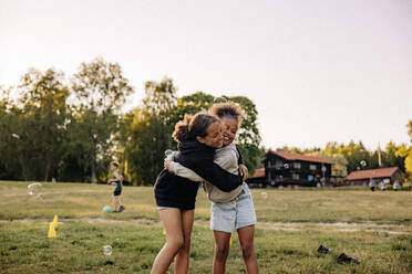 Glückliche Freundinnen umarmen sich gegenseitig, während sie auf dem Spielplatz im Gras stehen - MASF39507