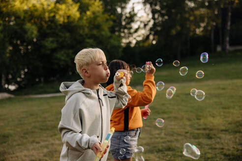 Boy blowing bubbles with female friend standing in playground at summer camp - MASF39511