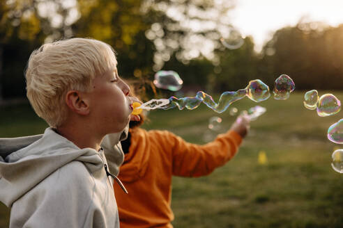 Side view of blond boy blowing bubbles while playing in playground - MASF39512