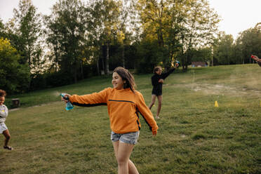 Smiling girl spraying water on friends while playing on grass in playground - MASF39519