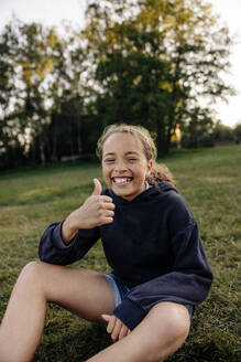 Porträt eines glücklichen Mädchens, das den Daumen nach oben zeigt, während es auf einem Spielplatz im Gras sitzt - MASF39540