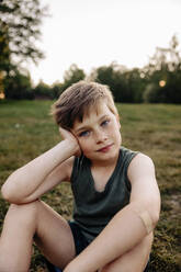 Porträt eines Jungen, der sich auf den Ellbogen stützt, während er auf einem Spielplatz im Gras sitzt - MASF39551