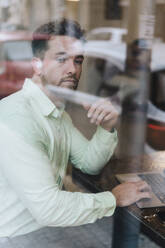 Thoughtful businessman sitting with laptop seen through glass in cafe - JOSEF20986