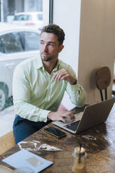 Thoughtful businessman sitting with laptop in cafe - JOSEF20987