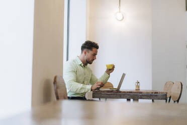 Geschäftsmann arbeitet an einem Laptop und hält eine Kaffeetasse in einem Cafe - JOSEF20989