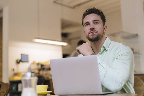 Thoughtful young businessman sitting with laptop at table in cafe - JOSEF20995