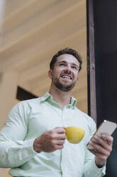 Glücklicher junger Geschäftsmann mit Kaffeetasse und Smartphone in der Tür eines Cafés - JOSEF20998