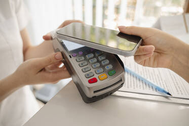 Hand einer Frau, die mit ihrem Smartphone auf einem Kreditkartenlesegerät in einer Klinik bezahlt - ALKF00605