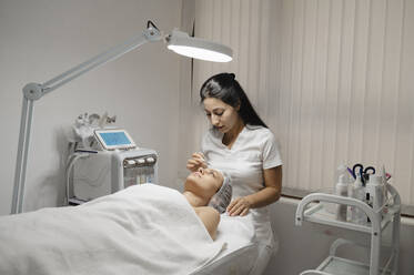 Dermatologe trägt in einer Klinik eine chemische Peeling-Maske auf das Gesicht einer Frau auf - ALKF00610