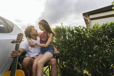 Smiling woman sitting on boyfriends lap near hedge - PBTF00239