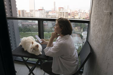 Frau sitzt mit Katze und verwendet Smartphone auf Balkon - YBF00227