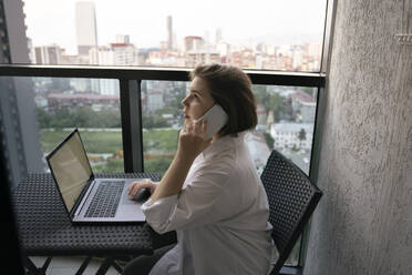 Young freelancer sitting with laptop and talking on smart phone on balcony - YBF00231