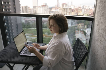 Freelancer sitting with laptop and using smart phone on balcony - YBF00232