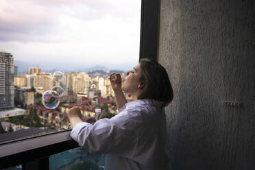 Junge Frau bläst Seifenblasen auf dem Balkon - YBF00233