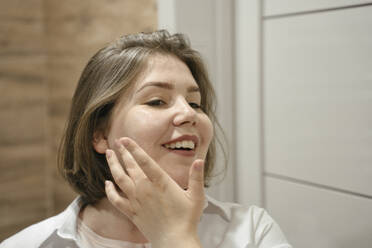 Lächelnde junge Frau trägt Feuchtigkeitscreme auf ihr Gesicht auf und schaut in den Spiegel - YBF00238