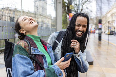 Fröhlicher Mann mit Frau, die ihr Smartphone in der Hand hält und auf dem Fußweg lacht - WPEF07660