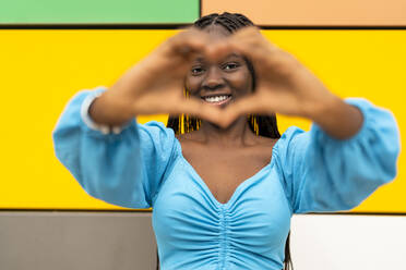 Smiling woman gesturing heart shape in front of multi colored wall - DLTSF03588