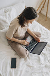 Freelancer sitting with legs crossed and using laptop on bed at home - EVKF00058